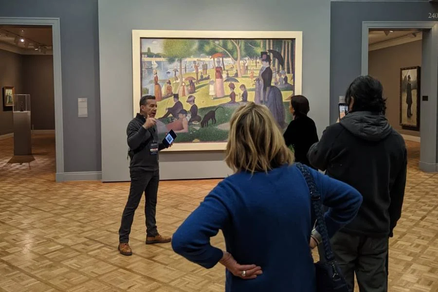 A museum guide speaks to a small group of visitors in front of Georges Seurat’s painting “A Sunday Afternoon on the Island of La Grande Jatte.”.